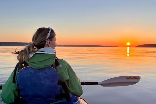 Person kayaking on calm water at sunset, wearing a green jacket and blue life vest.