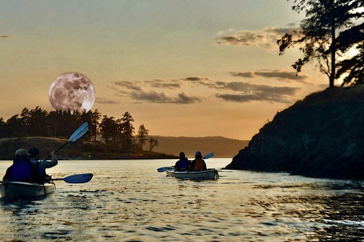 Kayakers on calm water with a large full moon in the background at sunset